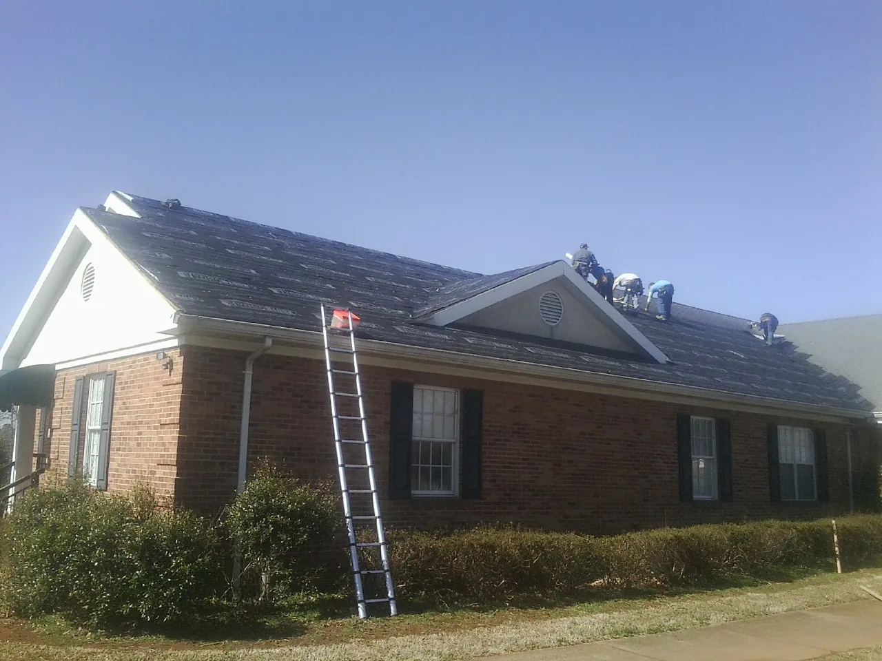 Crew tearing shingles off a brick ranch roof with a ladder set — Metal Roof Repair work by our crew in Saint Joe