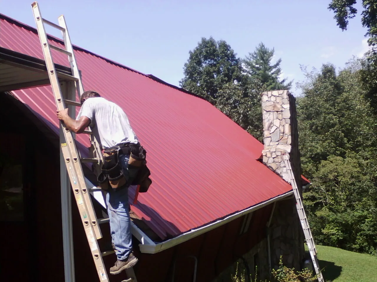 Roofer on a ladder beside a red metal roof and stone chimney — Soffit & Fascia Repair by Roofing Brothers in Saint Joe